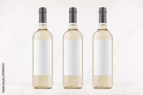 Three transparent wine bottles with blank white labels on white wooden board, mock up. Template for advertising, design, branding identity.