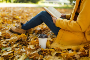Woman in yellow coat, jeans and boots sitting under the maple tree with a red book and cup of coffee or tea in fall city park on a warm day. Autumn golden leaves. Reading concept. Close up.