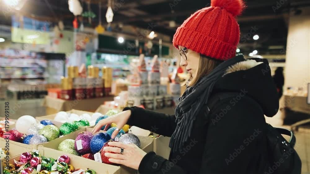 Young attractive caucasian girl with glasses and a red hat chooses the balls for the New Year tree in the market store. A woman is looking at colorful Christmas decorations. Young attractive caucasian girl with glasses and a red hat chooses the balls for the New Year tree in the market store. A woman is looking at colorful Christmas decorations.