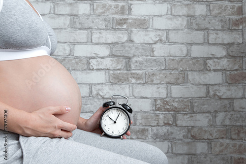 Pregnant woman holding an alarm clock while sitting on chair. Horizontal, copy space. Waiting for birth. Motherhood concept.