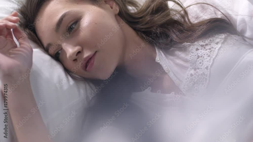 Slomo of beautiful woman in white nightgown lying in bed and looking sensualy into camera Slomo of beautiful woman in white nightgown lying in bed and looking sensualy into camera