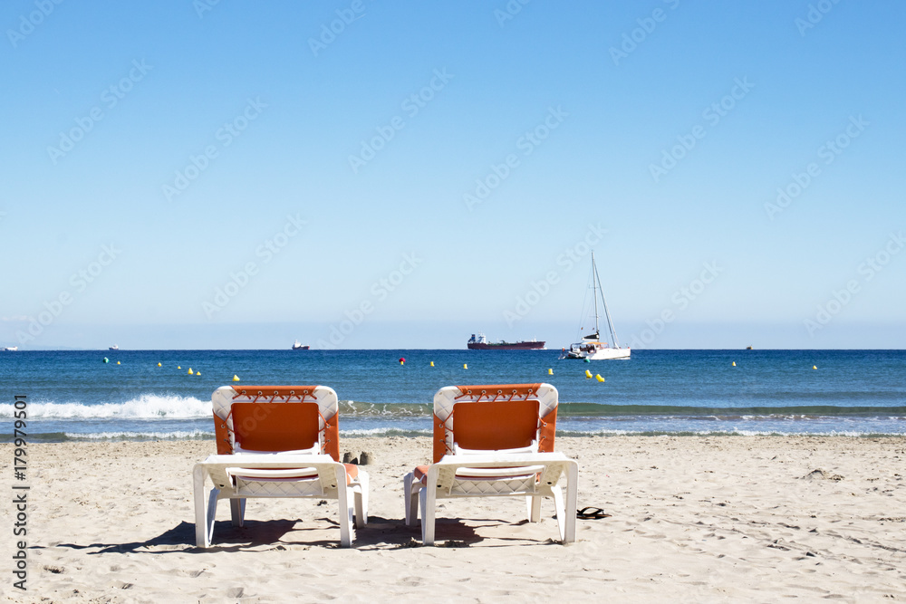 Two loungers on the beach are turned towards the sea with a view of yachts and ships.