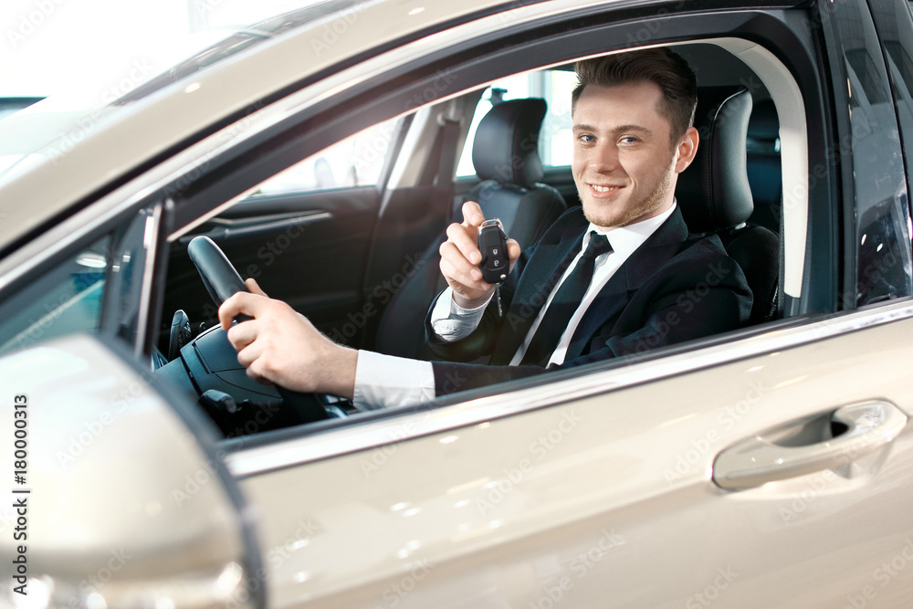 Formal wearing young man behind the wheel. He is looking at camera, showing the car key and smiling.