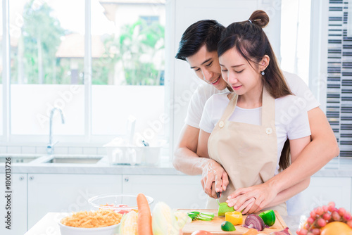 Asian lovers or couple cooking and slicing vegetable in kitchen room. Man and woman looking each other in home. Holiday and Honeymoon concept. Valentine day and wedding theme