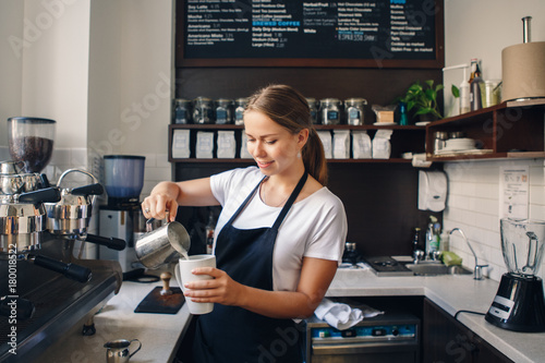 Portrait of smiling young Caucasian woman pouring hot milk in coffee. Waitress holding white mug cup in cafe. Person at work, small business concept