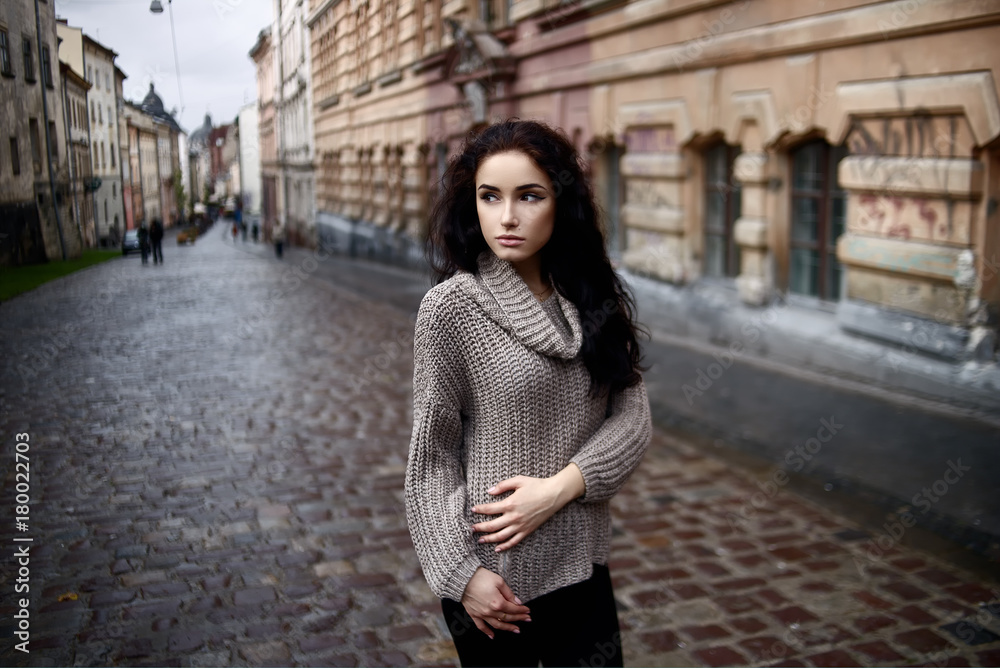 Portrait of romantic woman with long dark hair looking away in front of old wall. Outdoor photo of white girl in knitted gray sweater stands on the pavement. Copy space