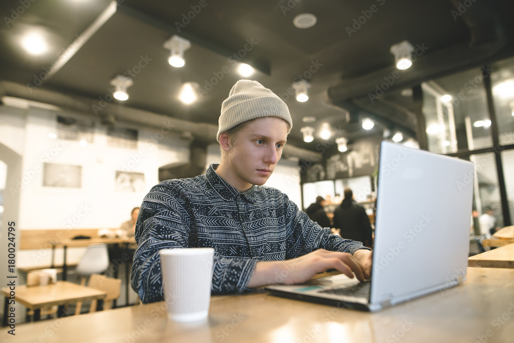 Fototapeta premium A hipster student sits in a cafe and works for a hand at a cup of coffee. Hipster sits in a cafe and uses a laptop.