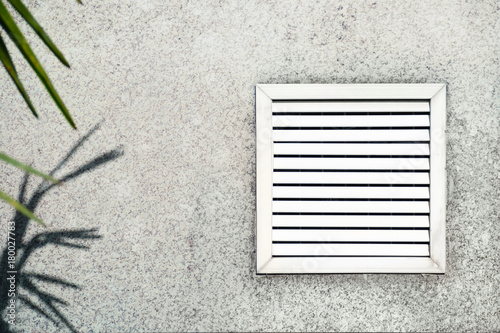 Aged vent with white shutters on the background of gray concrete under the leaves of the palm.