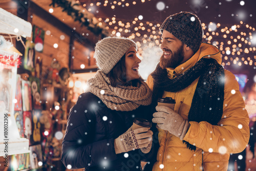 Having fun together at a christmas fairy with snowfall. Young cheerful couple is having a walk with hot drinks, enjoying, dressed warm, looking at each other and laugh, snowflakes all around