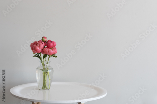 Small bunch of coral peonies in glass vase on round white table against neutral background with copy space