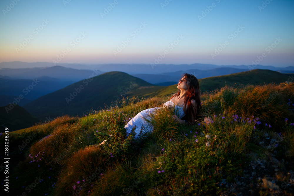 Beautiful woman in a long white dress in the mountains. Young woman sitting on a rock. Hair blowing in the wind. Girl relaxing outdoors. Beautiful woman posing in tall grass
