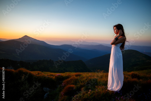 Young woman in long white dress. Beautiful young long hair woman posing in high mountain scenery. Woman standing on mountain top and contemplating the sunset