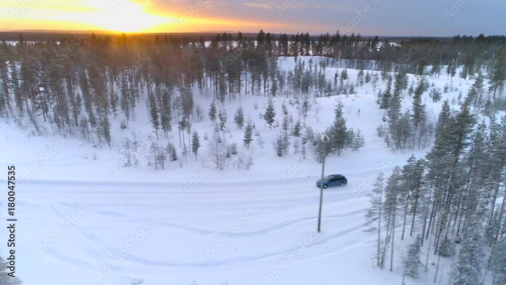 AERIAL Flying above blue car driving through snowy spruce forest at golden winter sunrise. People on road trip traveling across snow covered Lapland wilderness at sunset. Car driving on empty icy road AERIAL Flying above blue car driving through snowy spruce forest at golden winter sunrise. People on road trip traveling across snow covered Lapland wilderness at sunset. Car driving on empty icy road