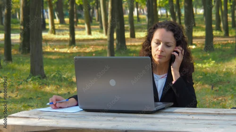 Business woman working on nature with laptop. Woman with laptop talking on the phone in the park. Business woman working on nature with laptop. Woman with laptop talking on the phone in the park.