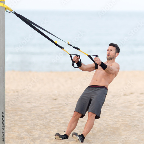 Athletic man making suspension training exercise on the beach 