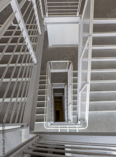 Looking down a rectangular staircase. You can see down three flights. No one is on the stairs. The stairs and railing are gray. You can barely see the yellow netting at the bottom of the stair well.