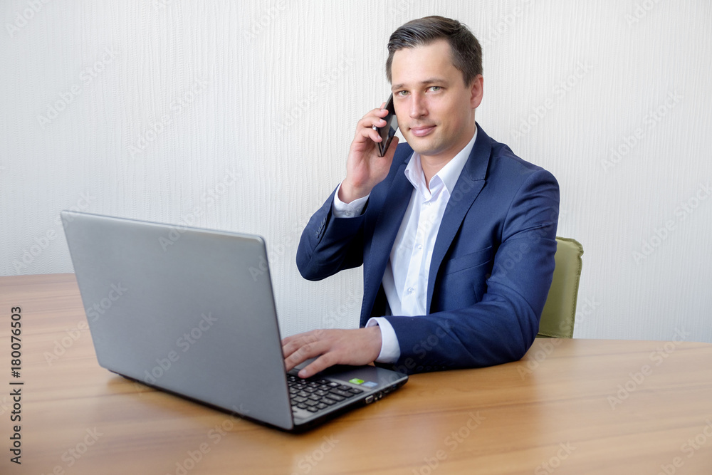 Handsome young concentrated attractive man using cellphone while working with laptop in the office