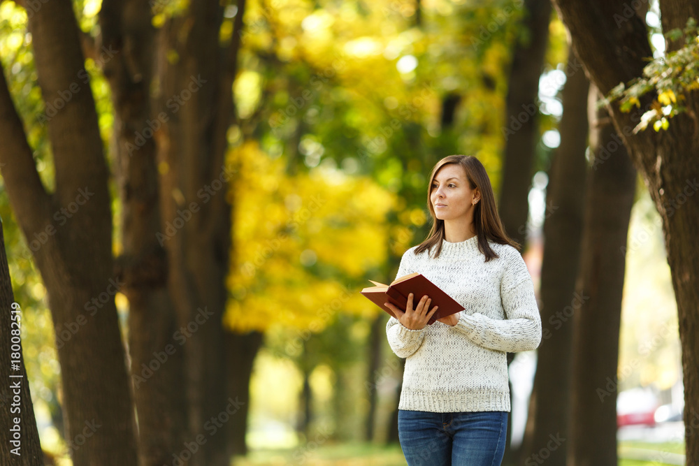 Naklejka premium A beautiful happy smiling brown-haired woman in white sweater standing with a red book in fall city park on a warm day. Autumn golden leaves. Reading concept.