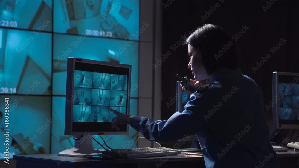 Single female security person monitoring closed circuit surveillance from multiple cameras in various rooms of a building informs on the radio to check the person who moves inside the room.