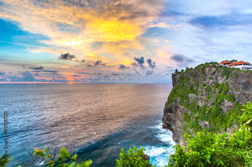 The Hindu temple on a high rock on the Indian Ocean in the southernmost part of the island against the backdrop of beautiful blue orange clouds at sunset. Pura Uluwatu temple, Bali, Indonesia.