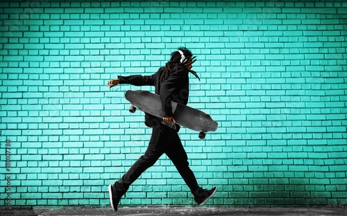 Handsome young dreadlocks skater with headphones running in a suit near the blue wall on the street.