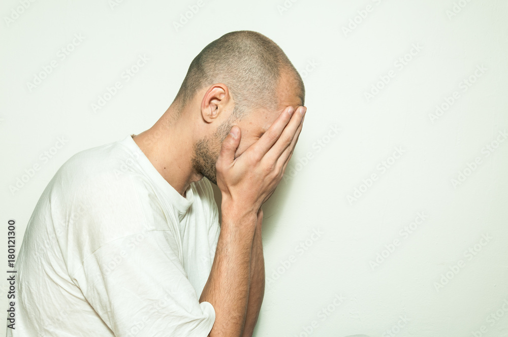 Fototapeta premium Young depressed man suffering from anxiety and feeling miserable cover his face with his hands and leaning on the white wall