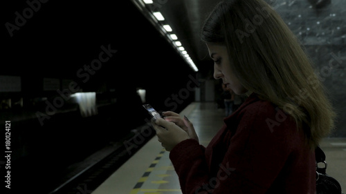 Girl in red coat using smartphone or tablet at subway station and waits for the train. Woman use of cellphone and standing at city subway staton.
