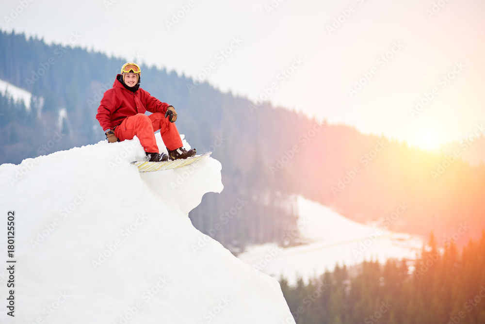 Young man snowboarder sitting on the top of the snowy hill with