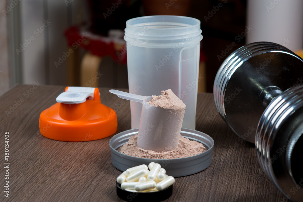 Whey protein in a measuring bucket with a metal dumbbell and shaker on a wooden background Whey protein in a measuring bucket with a metal dumbbell and shaker on a wooden background
