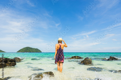 Girl in dress and hat walking into sea with blue clear water. Sky and island on background. Back view.
