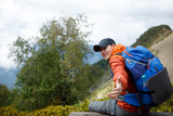 Image of sporty smiling man stretching out hand with backpack against backdrop of mountain