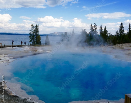Steaming, clear blue thermal pool beside lake with pine trees, blue sky, clouds and mountain in background