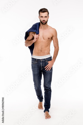 Stylish handsome. Full length of handsome young man in jeans looking at camera and holding his jacket while standing against white background.