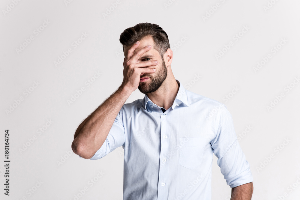 I can not look at it. Handsome young man looking at camera through his hand while standing against white background. I can not look at it. Handsome young man looking at camera through his hand while standing against white background.