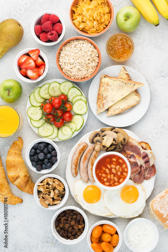 English breakfast with fried eggs sunny side up, sausages, bacon and mushrooms with fruits and vegetables, breads and juice on the grey white table, top view, vertical, selective focus © Liliya Trott
