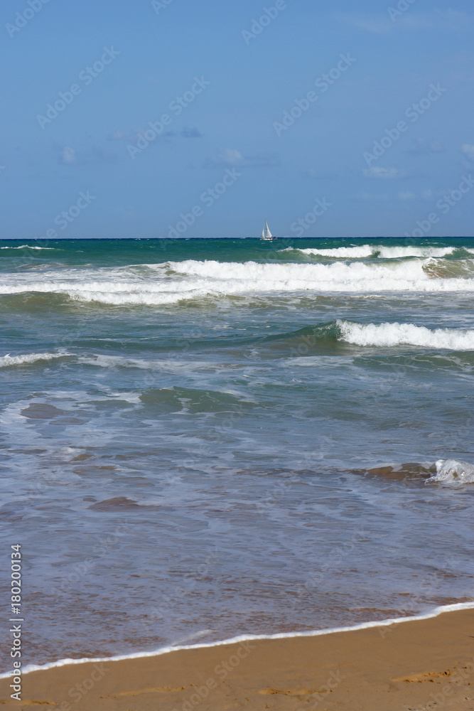 White sailboat on the horizon, sea waves at the sandy shore.