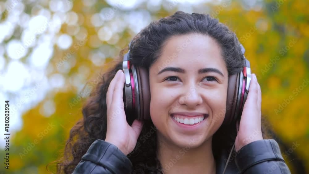 Close up portrait of latin curly haired girl listening music in headphones in autumn park. Beautiful young hispanic lady smiling, singing and enjoying melody outdoor. Slow motion. Close up portrait of latin curly haired girl listening music in headphones in autumn park. Beautiful young hispanic lady smiling, singing and enjoying melody outdoor. Slow motion.