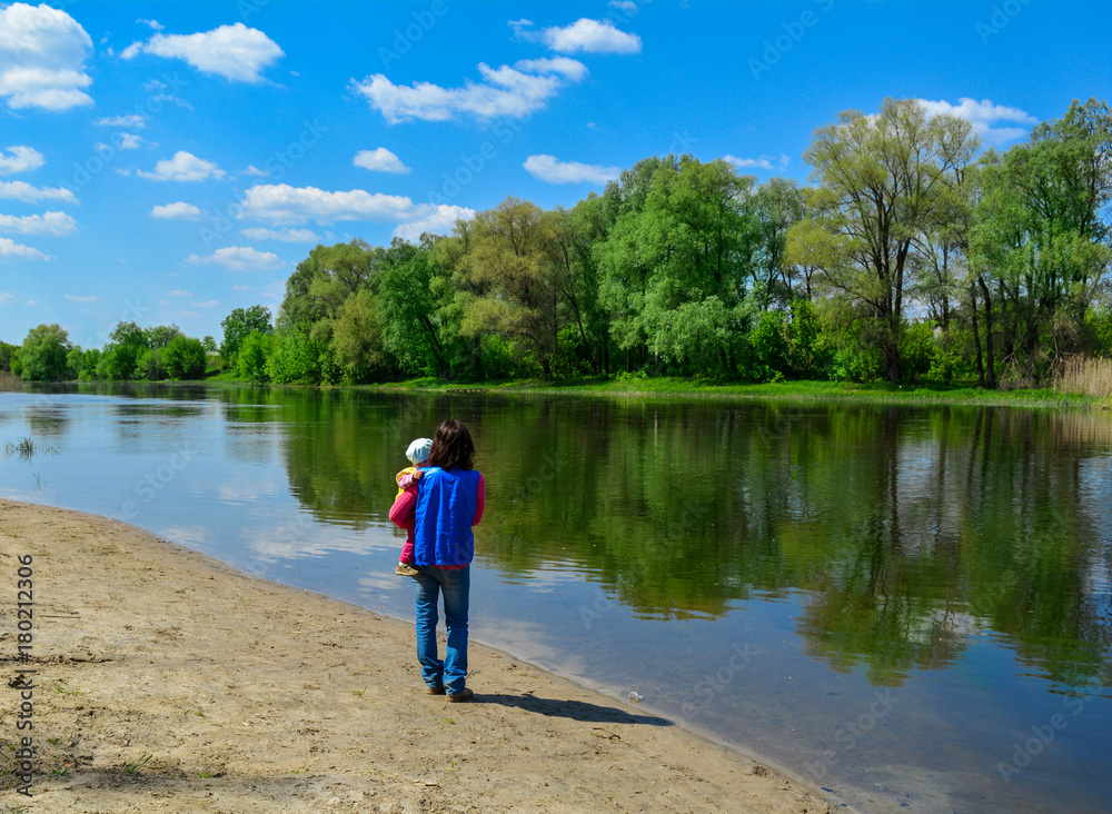 The young woman holds the child on hands and walks along the river in sunny spring day. The young woman holds the child on hands and walks along the river in sunny spring day.