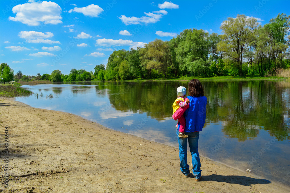 The young woman holds the child on hands and walks along the river in sunny spring day.