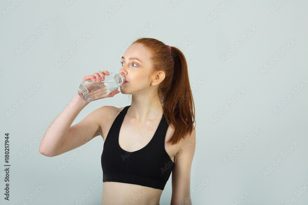 beautiful slender girl with red hair in sportswear on a light background with a bottle of water beautiful slender girl with red hair in sportswear on a light background with a bottle of water