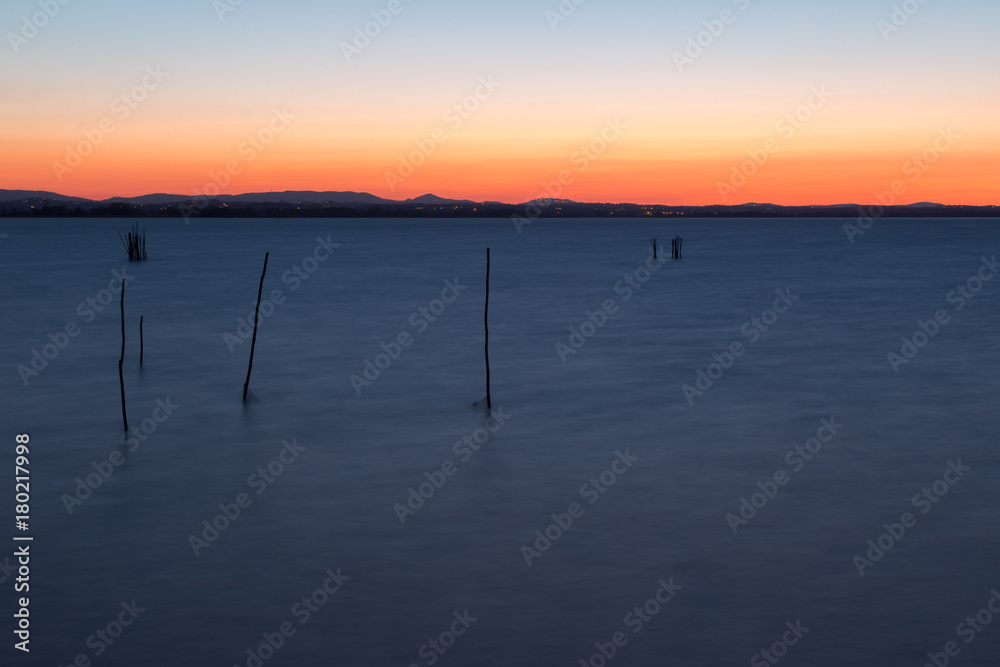 The end of sunset and the beginning of dusk on a lake, with colorful sky and dark, blue water, with poles and fishing nets