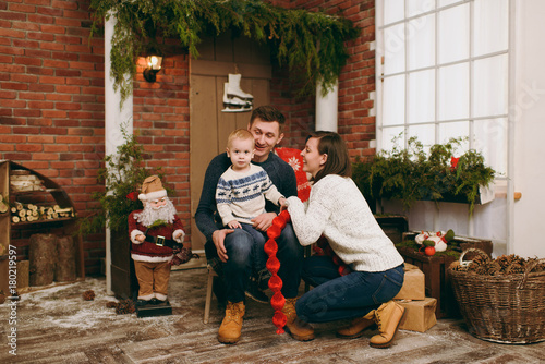 Young happy parents with a cute little child boy dressed in sweater sitting on the chair with Santa in decorated New Year room at home. Christmas good mood. Lifestyle, family and holiday 2018 concept © ViDi Studio