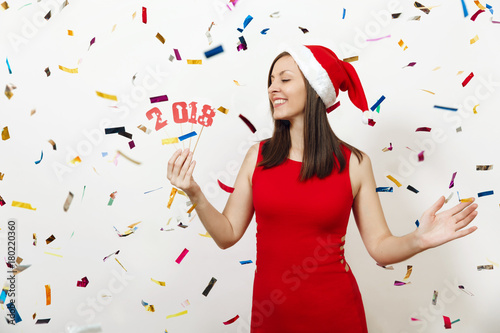 Beautiful caucasian young happy woman with healthy skin and charming smile in red dress and Christmas hat holding card number 2018 on white background. Santa girl isolated. New Year holiday concept