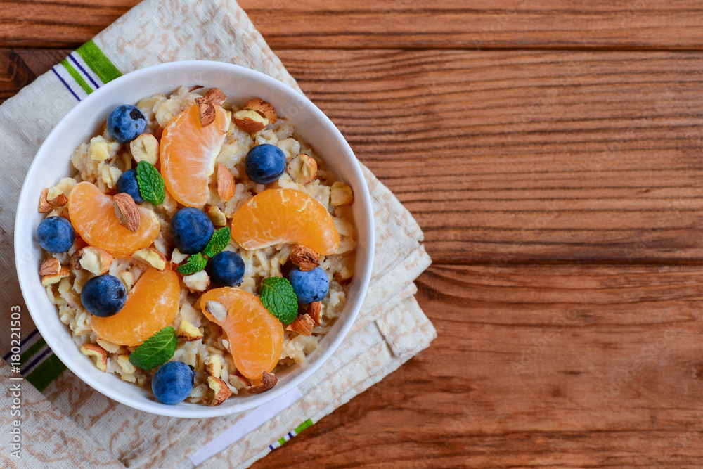 Light porridge with toppings. Oatmeal with juicy mandarins, blueberries, hazelnuts and mint in a white bowl and on a wooden background with copy space. Low calorie porridge recipe. Top view Light porridge with toppings. Oatmeal with juicy mandarins, blueberries, hazelnuts and mint in a white bowl and on a wooden background with copy space. Low calorie porridge recipe. Top view
