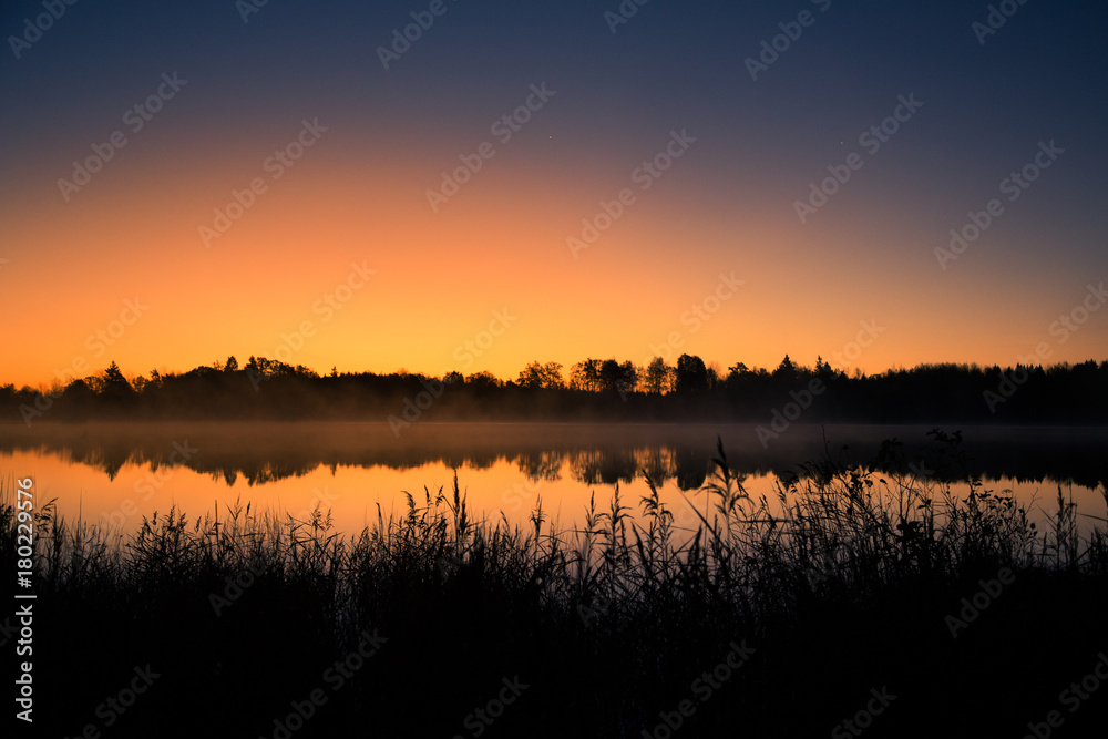 Obraz premium A beautiful sunrise on a lake with a raising mist. Beautiful morning in wetlands in Latvia. Autumn landscape in bright colors.