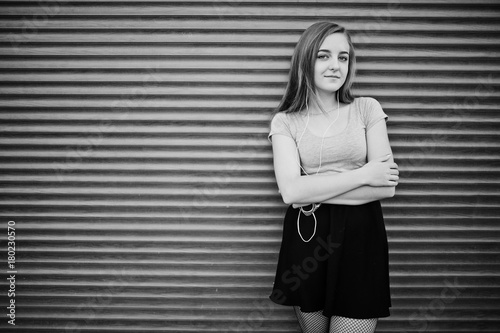 Young blonde girl in black skirt listening music from headphones of mobile phone against orange shutters.