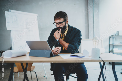 Young bearded male architect wearing eye glasses working on a digital tablet dock at his desk. Professional experienced engineer constructionist developer.Horizontal.Blurred background. photo