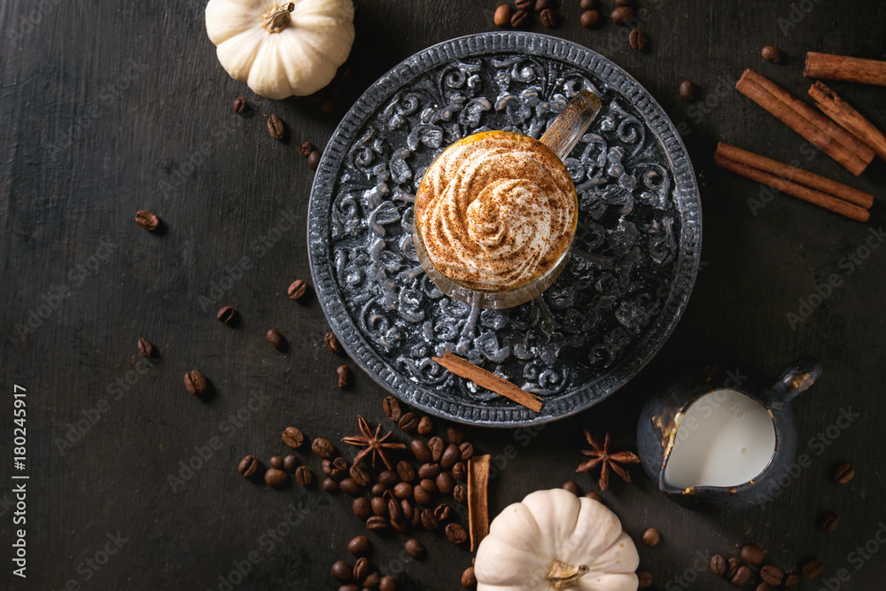Glass of spicy pumpkin latte with whipped cream and cinnamon standing on black ornate serving board with decorative white pumpkins . Coffee beans and spices above. Dark background. Top view, space