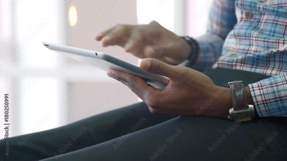 The man fills the form on his tablet while sitting in bright room. Male holds his brand-new device by his left hand with stylish wristwatches on and scroll down the page by the right one. The person