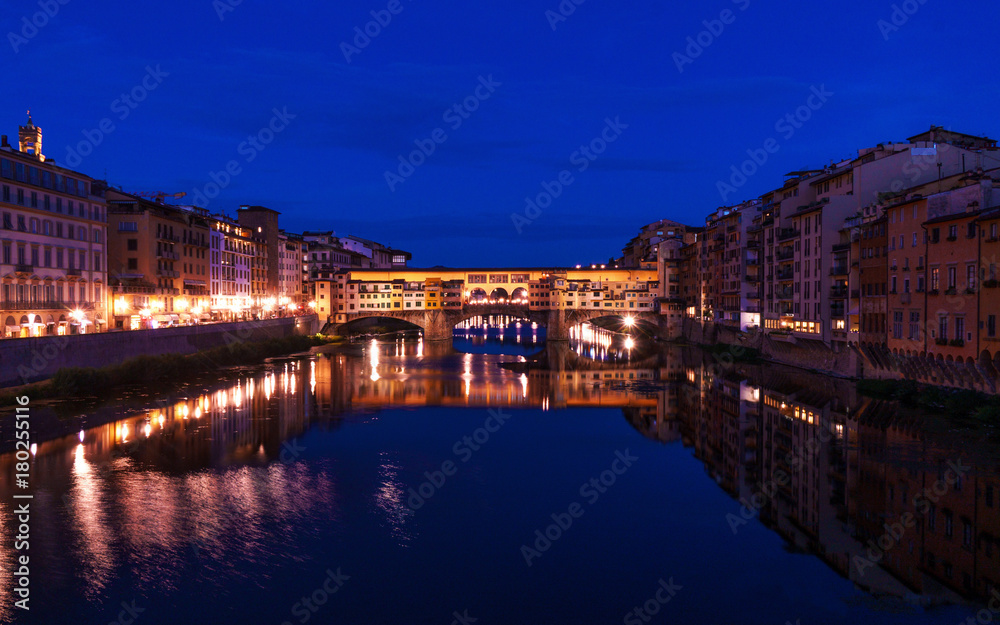 Ponte Vecchio bridge in Florence at night time with city lights reflecting in Arno river.